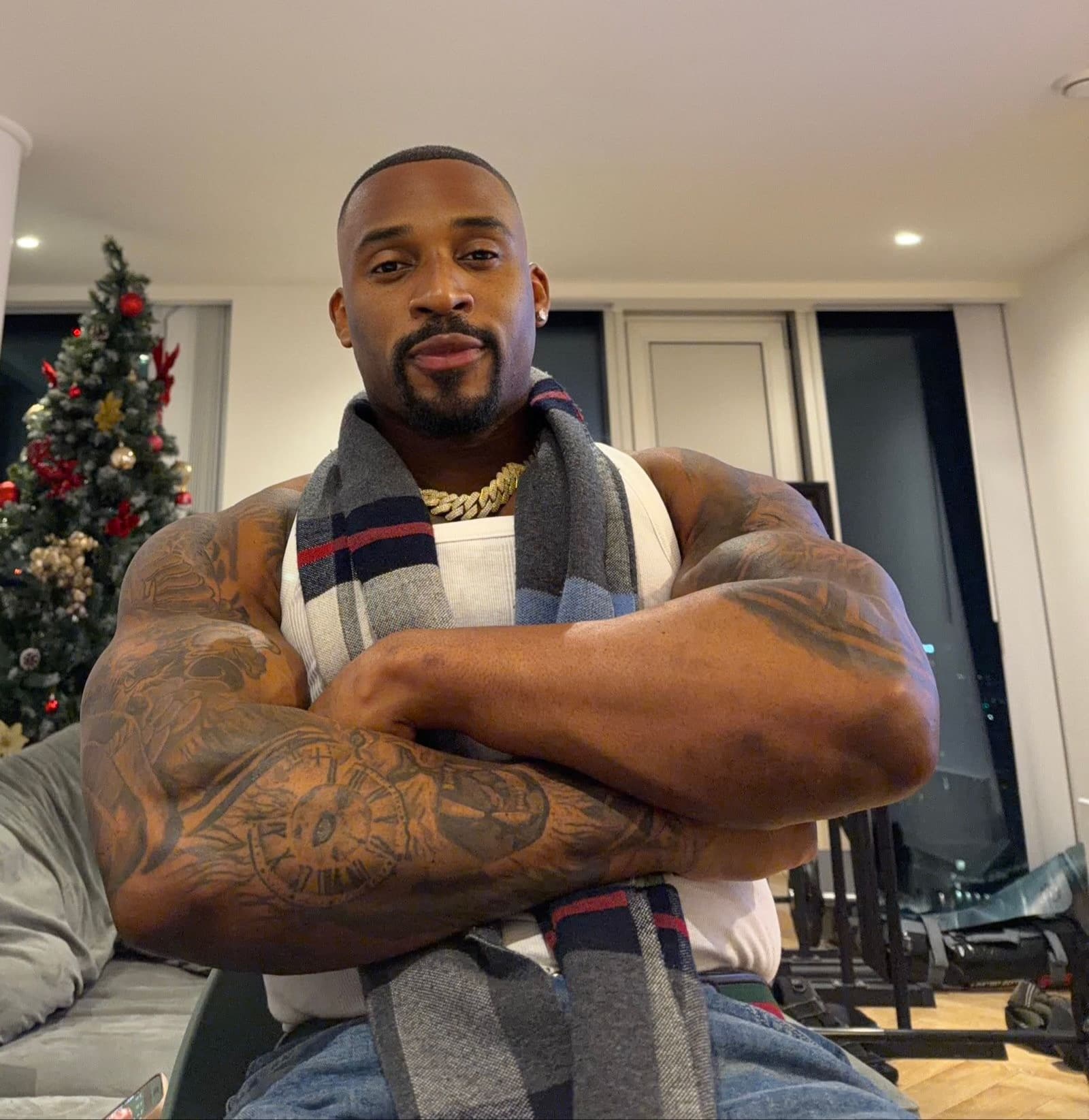 Tattooed muscular man in tank top and scarf, arms crossed, with a Christmas tree behind.